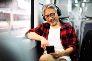 Hörbuch-Apps im Test: Ein älterer Herr sitzt im Zug und schaut auf sein Smartphone, während er Kopfhörer trägt.