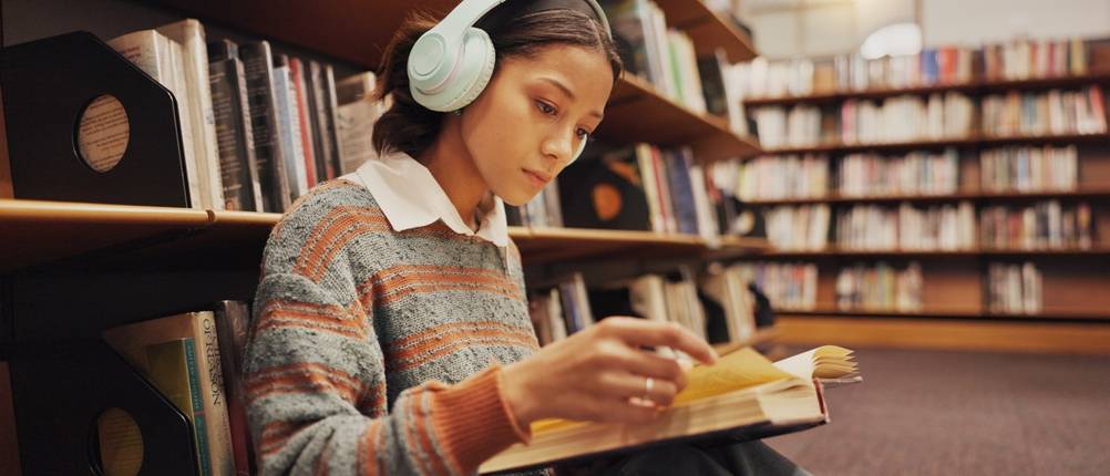 Junge Frau mit Kopfhörern liegt vor einem Bücherregal und liest in einem aufgeschlagenen Buch