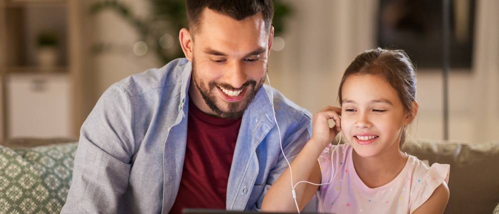 Vater und Tochter sitzen zu Hause auf dem Sofa vor einem Tablet und teilen sich ein Paar In-Ear-Kopfhörer