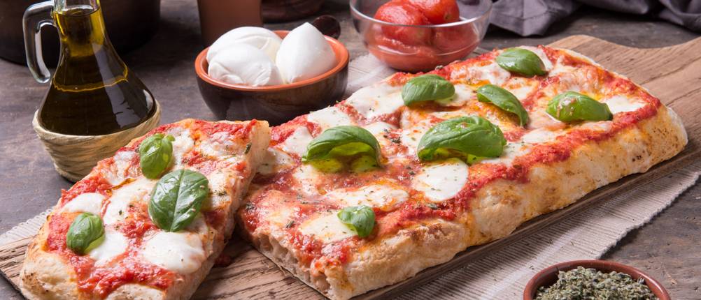 Rechteckige Pizza mit Mozzarella und Basilikum liegt auf einem Holzbrett. Daneben stehen Öl und Schälchen mit Tomaten, Mozzarella und Kräutern.