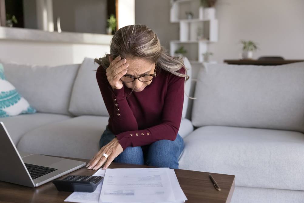 EIne Frau sitzt auf dem Sofa und hält sich verzweifelt die Hand an den Kopf. Vor ihr liegen Rechnungen, ein Taschenrechner und ein Laptop.