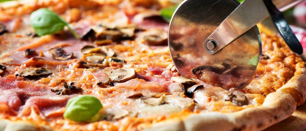Pizza mit Schinken und Pilzen wird auf einem Holzbrett mit einem Edelstahl-Pizzaschneider geschnitten.