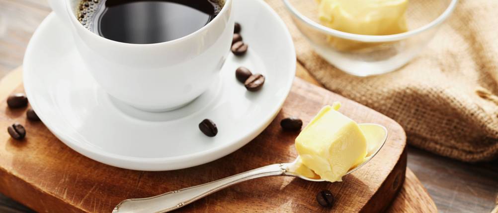 Auf zwei gestapelten Holzbrettchen sind eine Tasse Kaffee, ein Teelöffel mit Butter und Kaffeebohnen platziert.