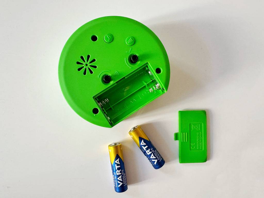 Im Test: Ein grüner Kinderwecker liegt mit der Rückseite nach oben, geöffnetem Batteriefach und 2 Batterien und Deckel nebenliegend auf einer weißen Fläche.