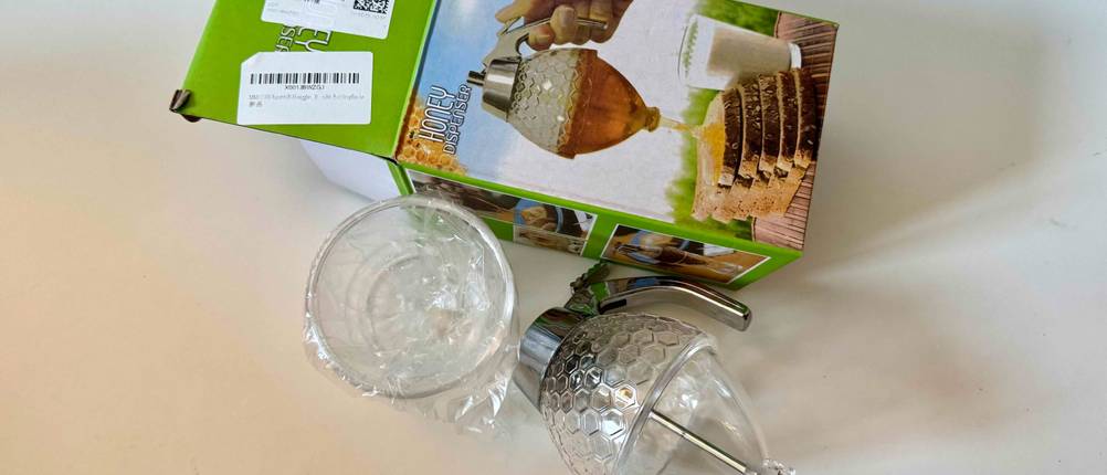 Neben der Originalverpackung liegt ein geöffneter und getesteter Honigspender auf einer hellen Fläche.