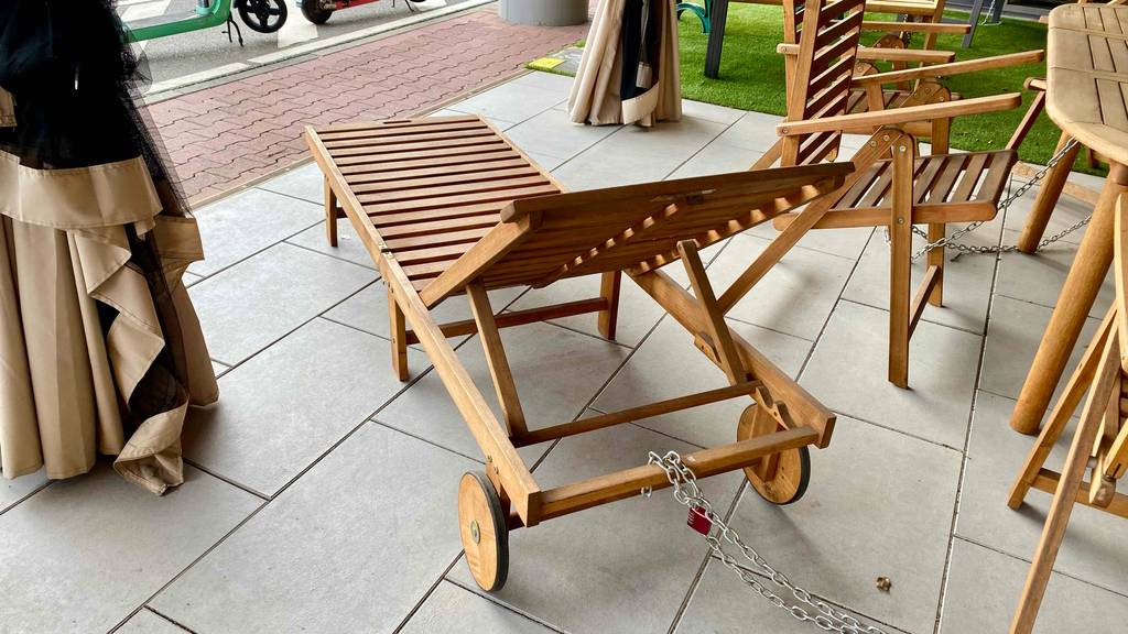 Ansicht von hinten auf den Verstellmechanismus des Kopfteils einer Holz-Gartenliege im Test, die auf einem hellgrauen Fliesenbelag im Geschäft neben anderen Gartenmöbeln steht.