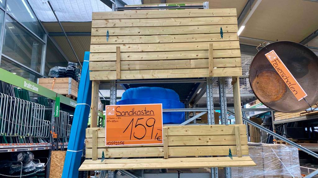 Ansicht von vorn auf einen an der Regal-Stirnseite befestigten, hellen Sandkasten aus Holz im Test, der in einem Baumarkt ausgestellt ist.