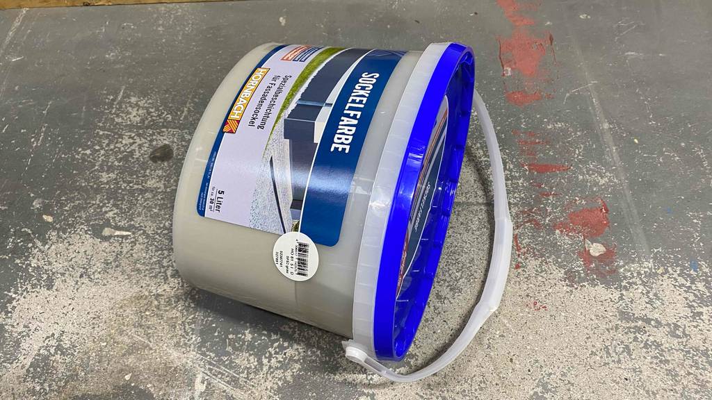 Ein Eimer getestete Sockelfarbe liegt quer auf einem Betonboden.