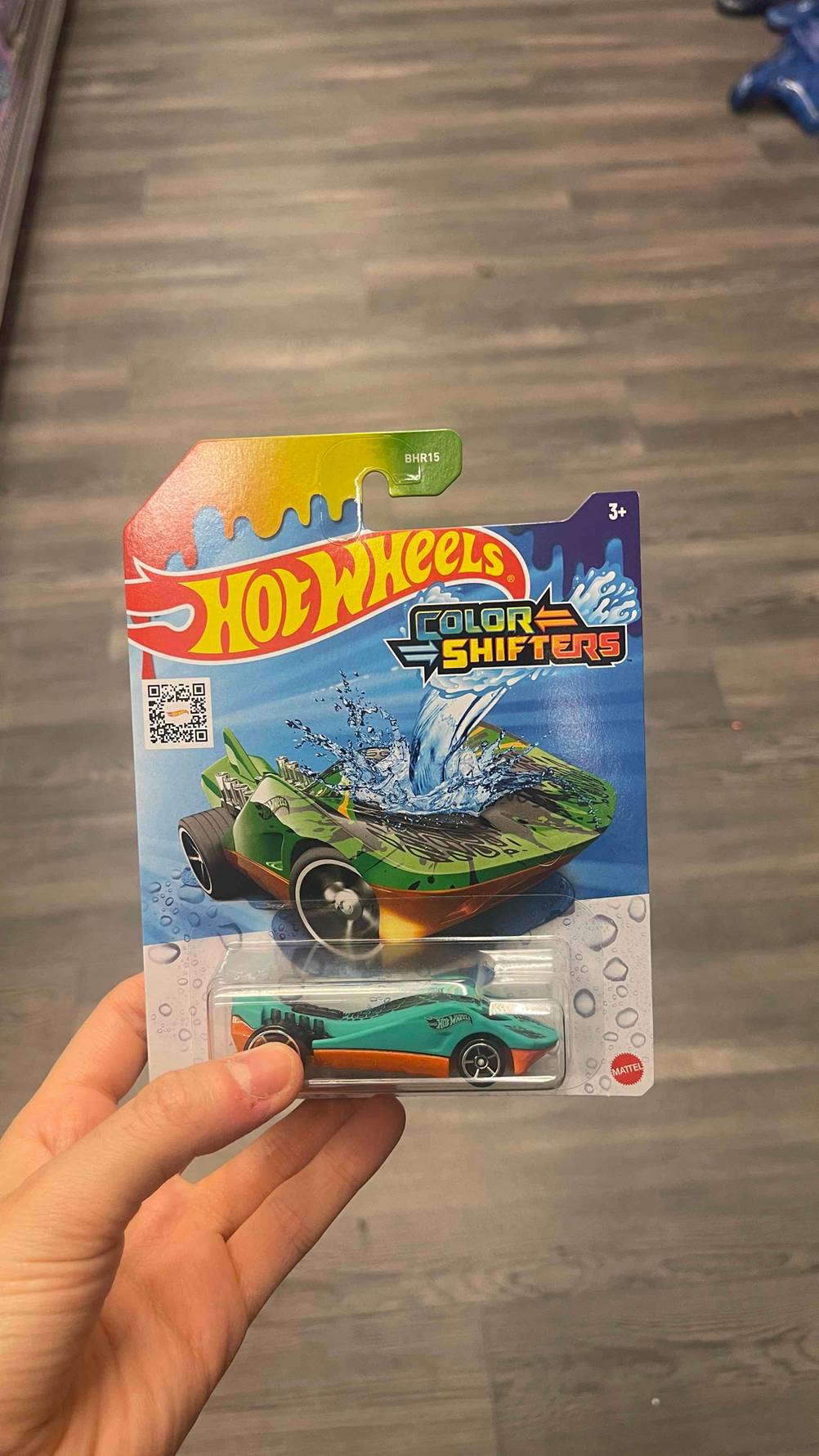 Hot-Wheels-Autos-Test: Eine Hand hält eine Verpackung mit einem grün-orangenen Modell über einem grauen Boden.