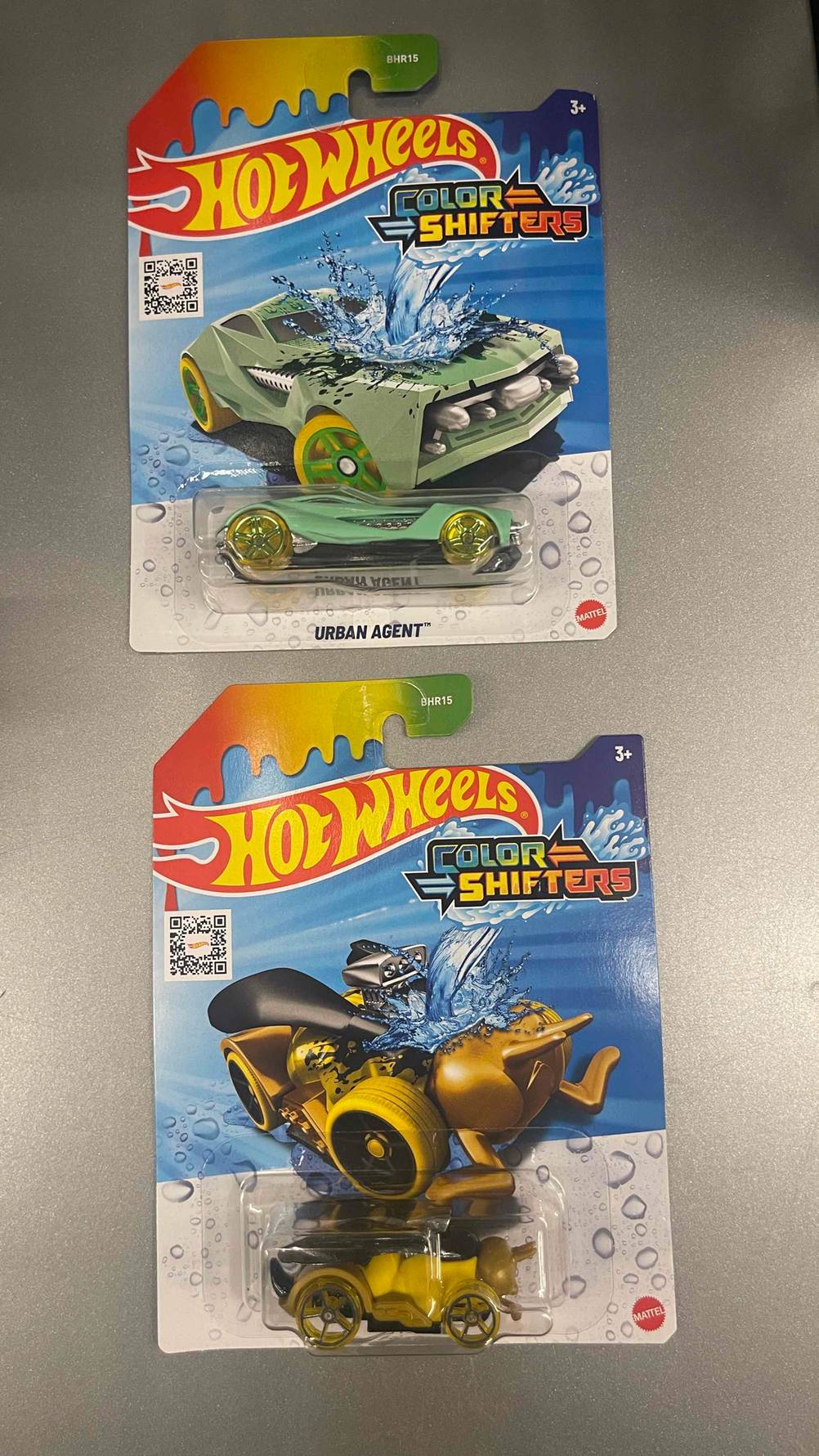 Getestet: Zwei Hot-Wheels-Autos in unterschiedlichem Design liegen innerhalb ihrer Verpackungen auf einer grauen Oberfläche.