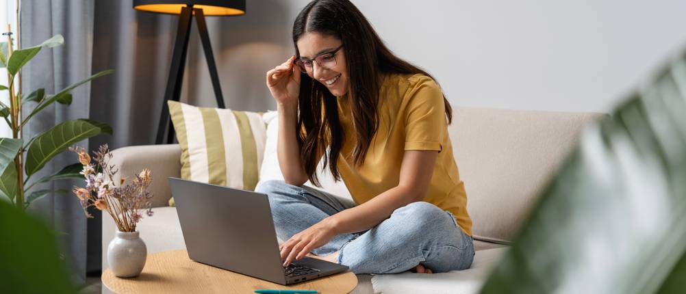 Eine Frau sitzt auf ihrer Couch und schaut glücklich auf den Bildschirm ihres Laptops.
