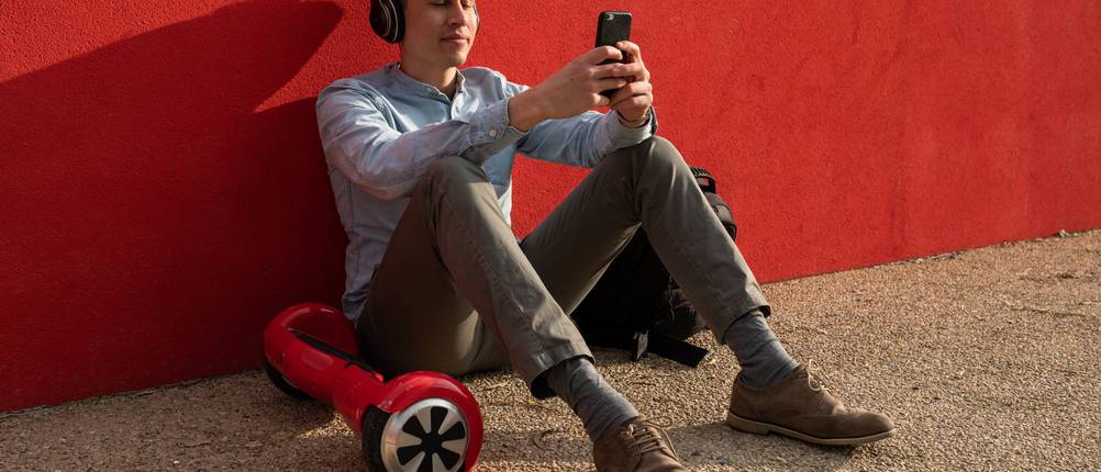 Hoverboard-Test: jungerr Mann mit lächelndem Gesicht, der neben seinem Hoverboard auf dem Asphalt sitzt und sein Smartphone mit Kopfhörern benutzt