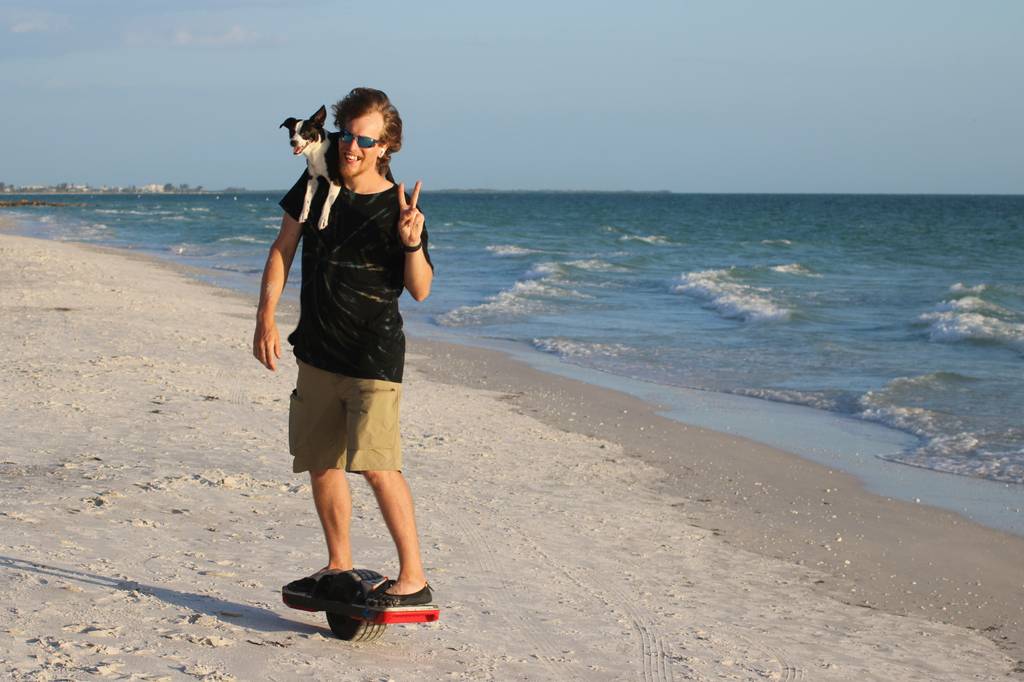 Hoverboard-Test: junger Mann fährt mit seinem Hoverboard am Strand entlang, auf der Schulter sitzt sein Hund