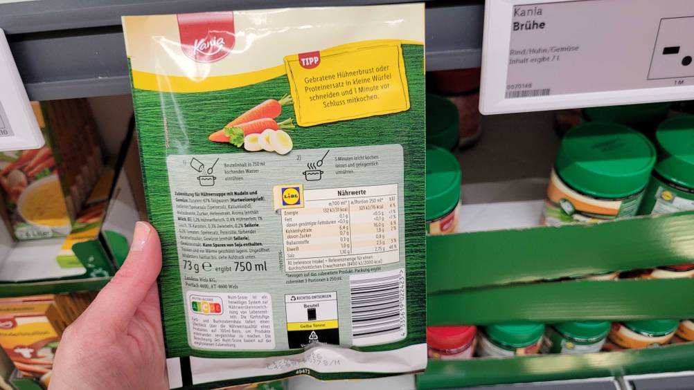 Die Rückseite einer Packung der getesteten Hühnersuppe von einer Hand vor ein Regal gehalten.