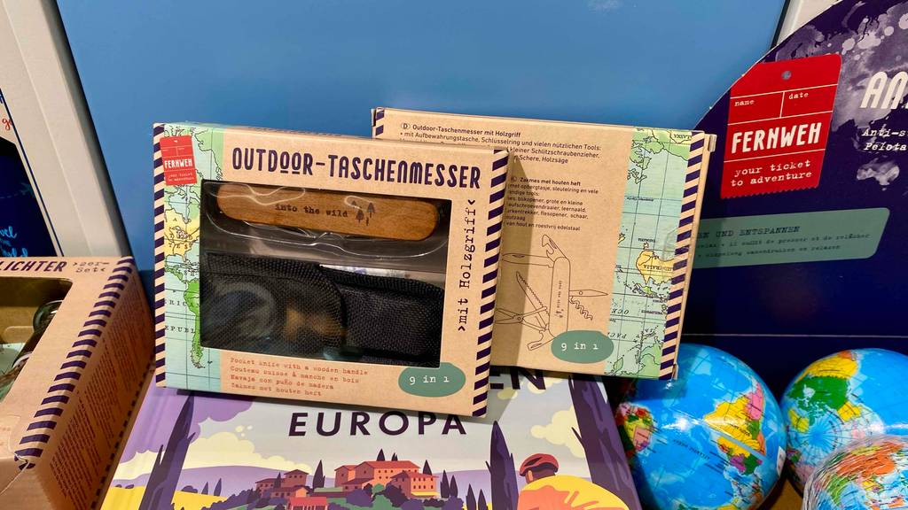 Im Test: Zwei verpackte Outdoormesser einmal die Vorderseite einmal die Rückseite vor einem blauen Hintergrund auf anderen Produkten stehend.