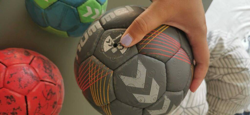 Ein getesteter Handball in Grau befindet sich in einer Damenhand, im Hintergrund sind weitere Bälle zu sehen.