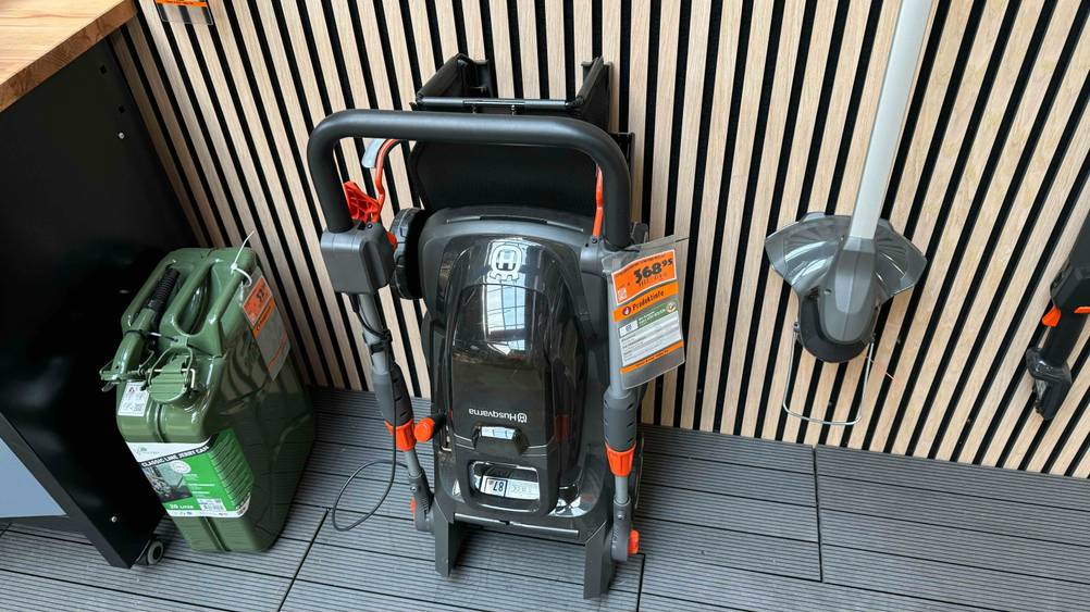 Ein getesteter Husqvarna-Rasenmäher mit Preisschild steht vor einer Holzwand auf einem dunklen Boden.
