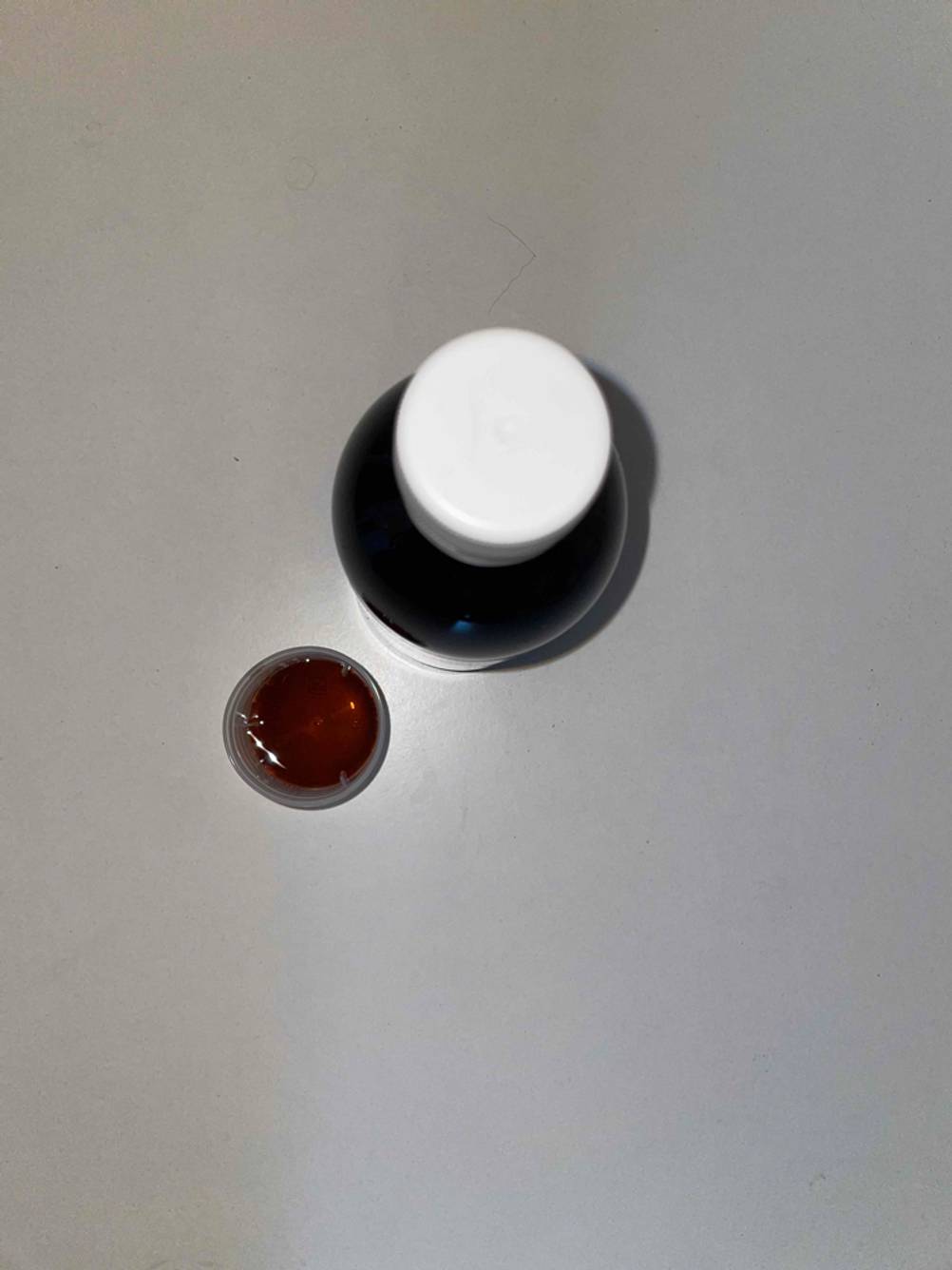 Auf dem Boden steht eine Glasflasche mit getestetem Hustensaft neben einer Dosierhilfe. 