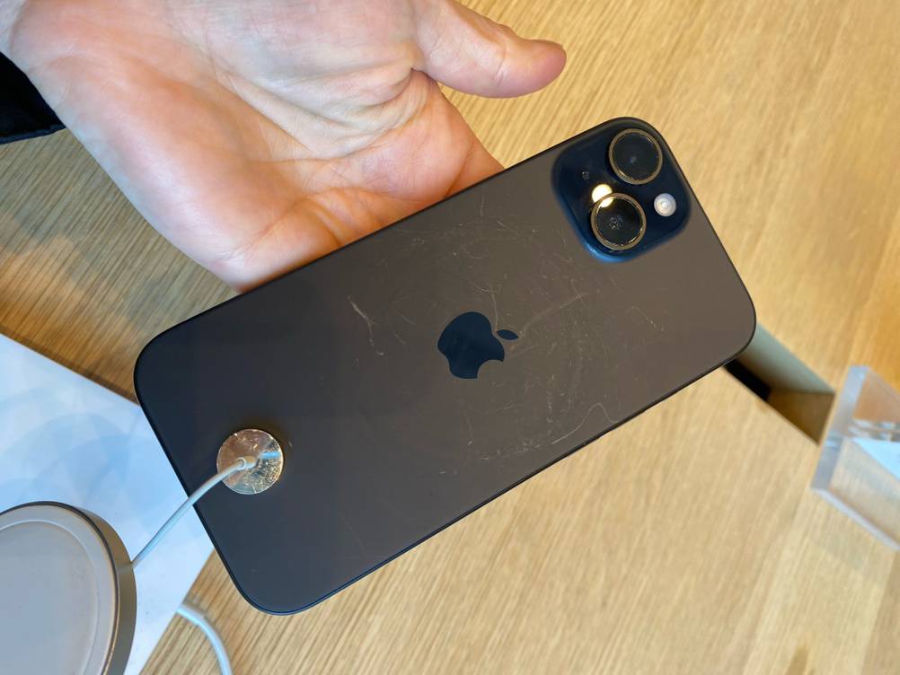 Hand hält ein gesichertes iPhone Handy mit guter Kamera im Test, den Fokus auf die matt schwarze Rückseite gerichtet, über einem Ausstellungstisch aus Holz.