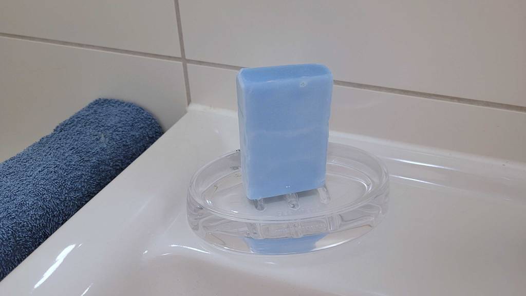 Seifenschalen im Test: Eine transparente Seifenschale inklusive hellblauer Seife steht auf einem weißen Waschbeckenrand neben einem Handtuchhalter mit blauem Frotteehandtuch.