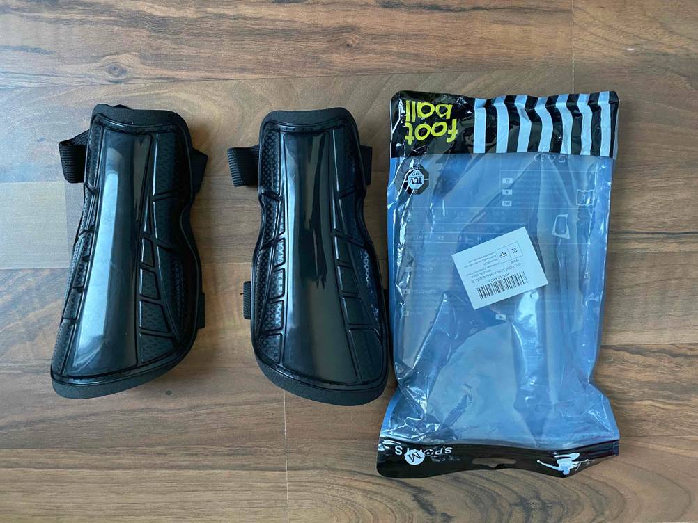 Zwei schwarze Schienbeinschoner im Test auf Holzboden, mit Fokus auf äußere Hartschalung und dazugehöriger Verpackung.