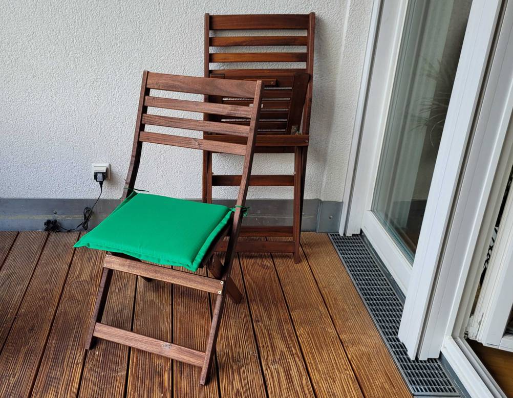 Gartenstühle-Test: Ein Gartenstuhl lehnt zusammengeklappt an einer Balkonwand, davor steht ein weiter Holz-Gartenstuhl aufgeklappt mit grüner Sitzauflage.