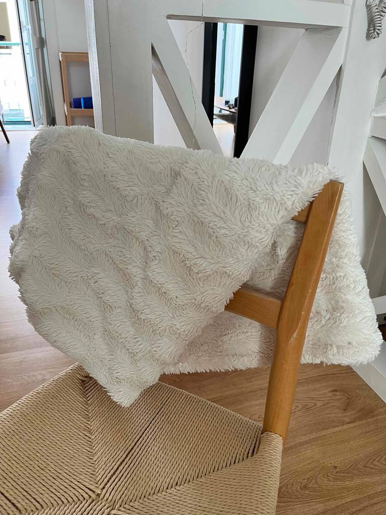 Eine weiße getestete Kuscheldecke in Langhaaroptik liegt zusammengefaltet über der Rückenlehne eines Holzstuhls im Wohnzimmerbereich.