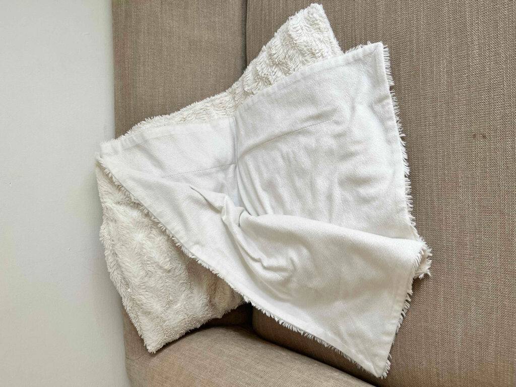 Ansicht auf die plüschige Innenseite einer weißen, getesteten Kuscheldecke, die auf einem beigefarbenen Sofa liegt.