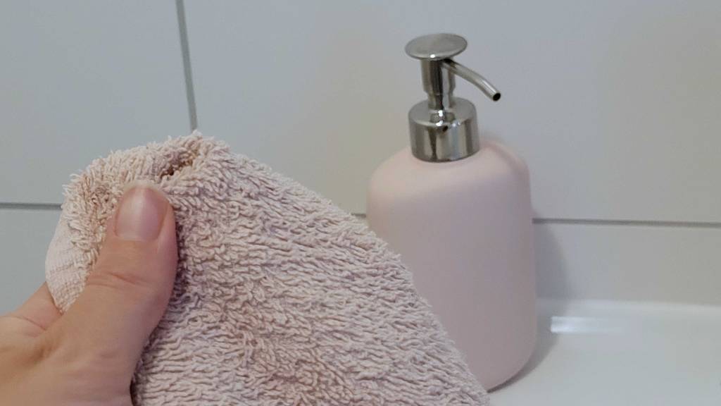 Hand hält ein Frottee-Handtuch in Altrosa vor einem rosafarbenen, stehenden Seifenspender im Test, der mit einem silberfarbigen Pumpmechanismus ausgestattet ist.