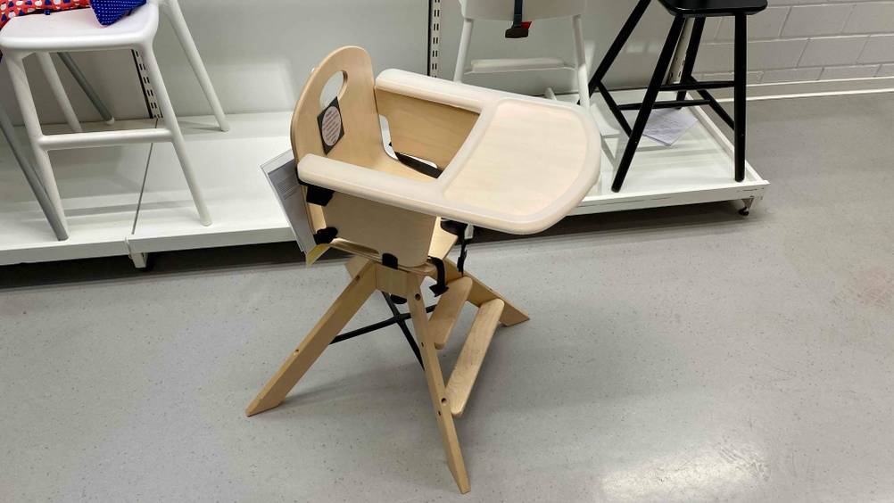 Hochstuhl getestet: Ein Produkt von schräg oben auf einem grauen Boden, im Hintergrund auf einem Sockel andere Produkte.