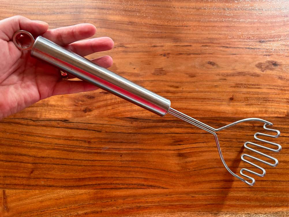 Hand hält einen getesteten Kartoffelstampfer über Holzfläche.