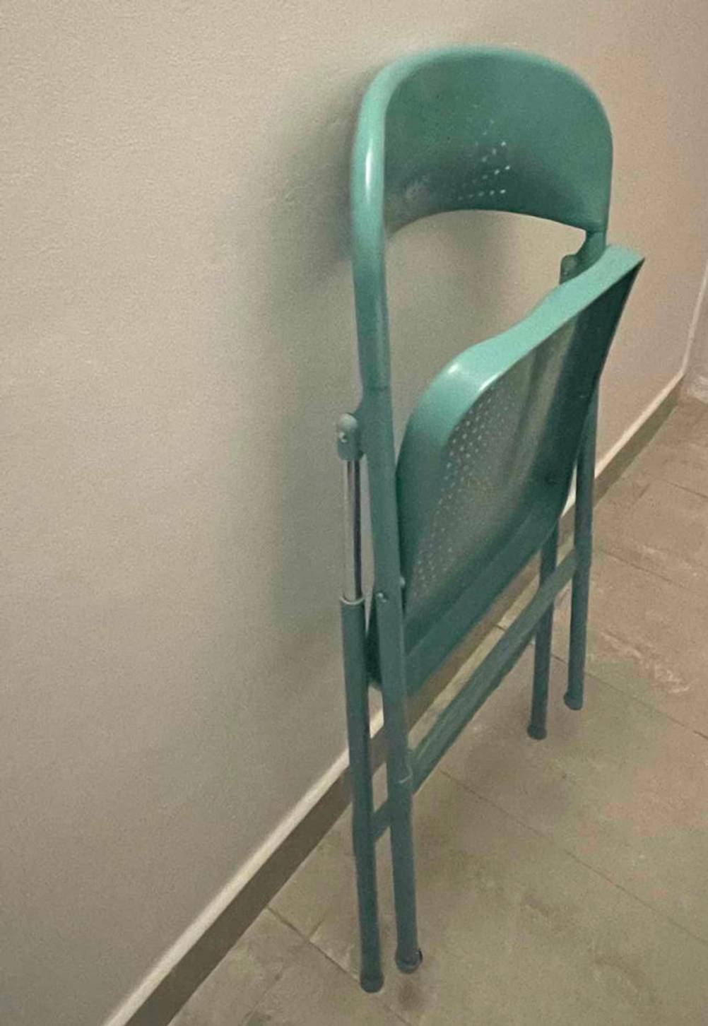 Seitenansicht eines hellgrünen Klappstuhls im Test im Loch-Design, der zusammengeklappt an einer weißen Wand steht.