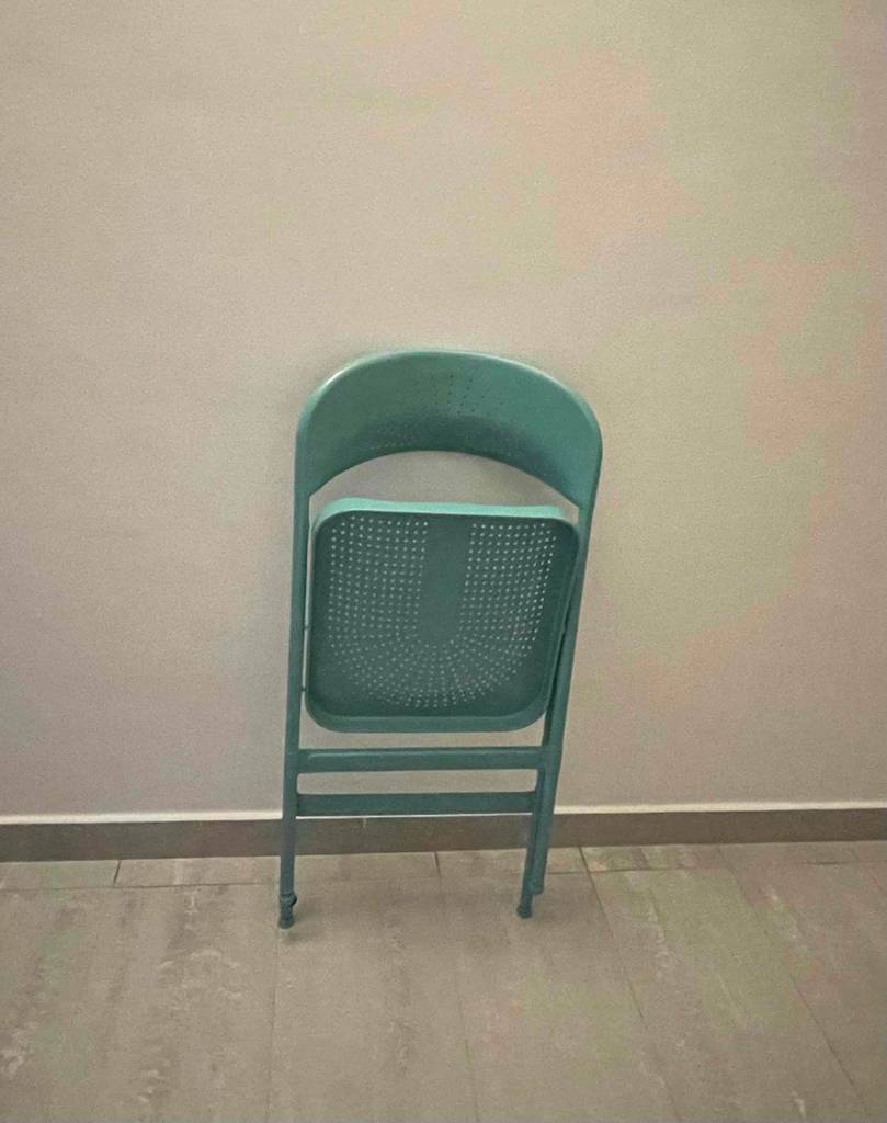 Ansicht von vorn auf einen zusammengeklappten und getesteten Klappstuhl in Grün, der an einer weißen Wand lehnt.