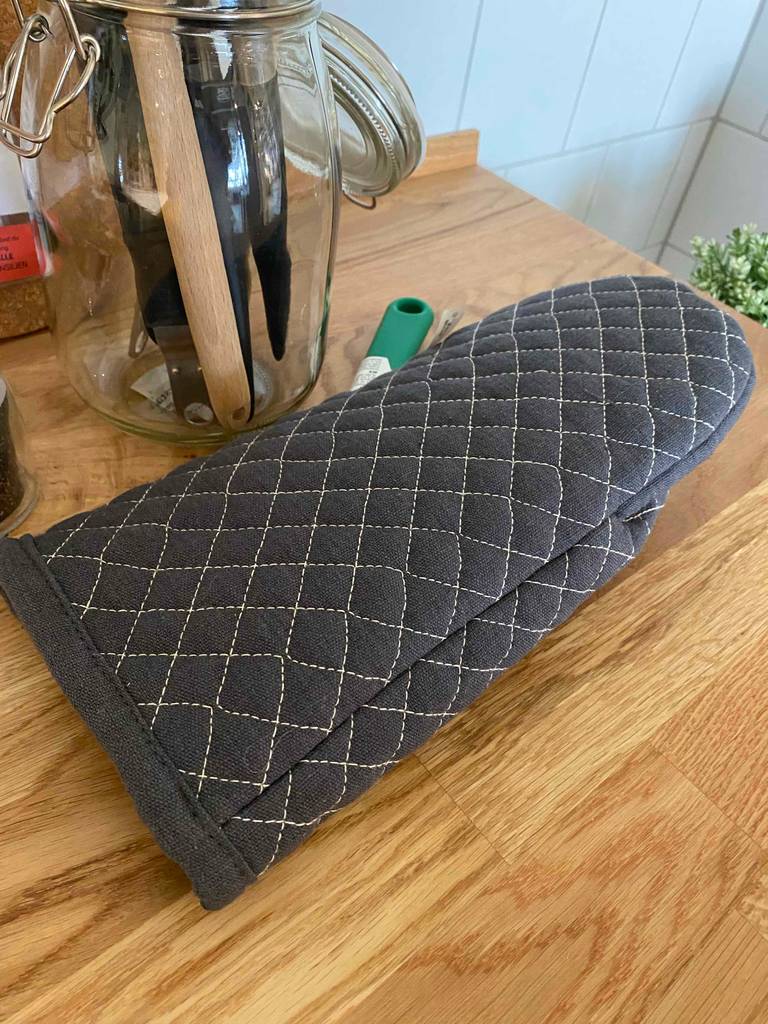 Ein grauer getesteter Ofenhandschuh liegt auf einem Küchentisch vor einem Glas mit Kochutensilien.