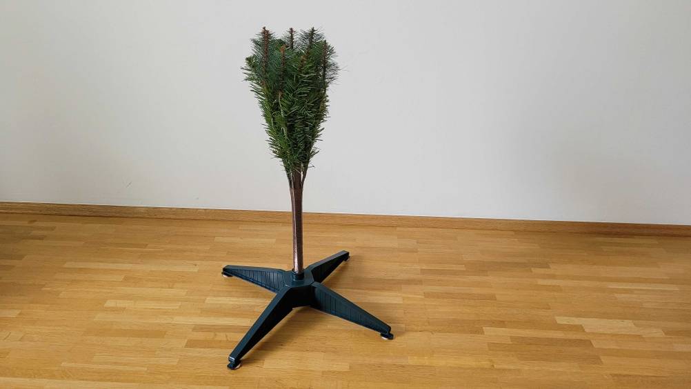 Ein getesteter künstlicher Weihnachtsbaum steht halb auseinandergefaltet in dem aufgebauten Ständer auf einem Holzboden.
