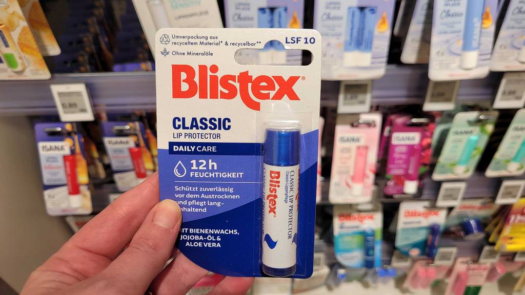 Eine Hand hält eine original verpackte Blistex-Lippenpflege im Test vor einem Drogeriemarktregal.
