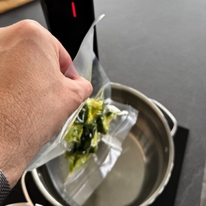 Eine Hand legt vakuumierten Brokkoli in einen Kochtopf mit Wasser