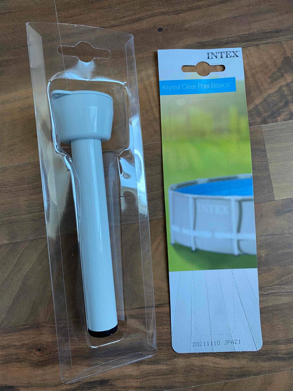 Weißes, analoges, getestetes Poolthermometer befindet sich in einer geöffneten Originalverpackung auf einer Holzfläche.