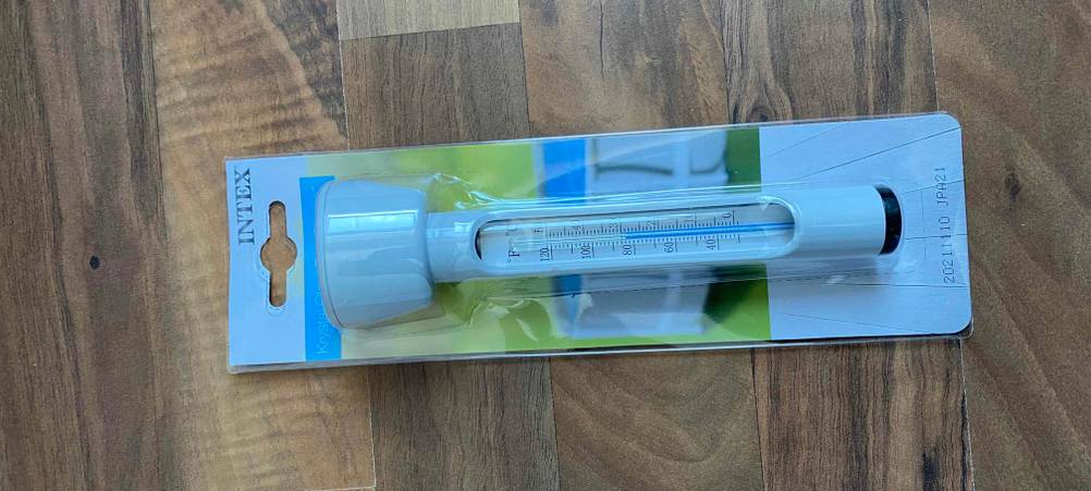 Weißes, analoges Intex Poolthermometer im Test liegt in seiner Originalverpackung auf einem Holzboden.
