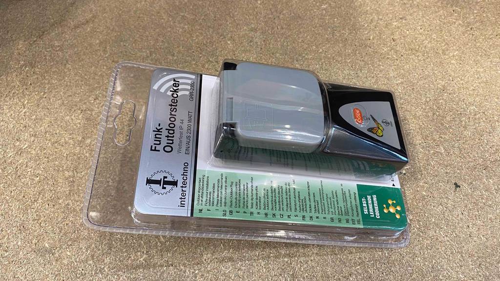 Außen-Funksteckdose-Test: transparente Kunststoffverpackung mit einem Funk-Outdoorstecker von Intertechno liegt auf einem braunen Untergrund.