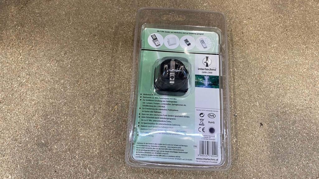 Funksteckdose für außen getestet: Eine transparente Kunststoffverpackung mit der Rückseite nach oben inklusive Funk-Outdoorstecker liegt auf einem grauen Boden.