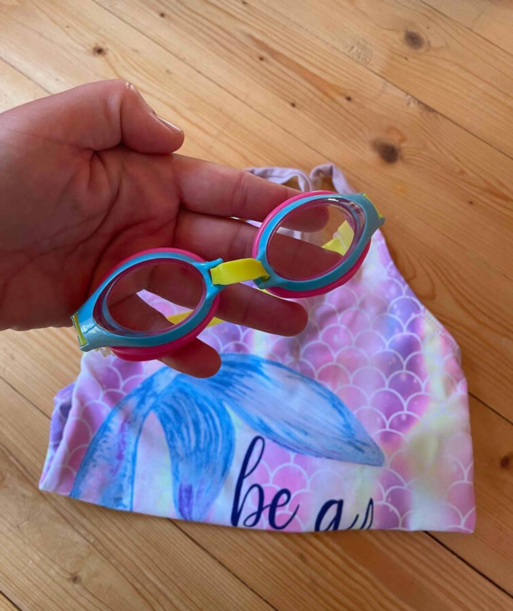 Eine Hand hält die getestete Schwimmbrille für Kinder über einem Kinder-Badeanzug in Rosa.