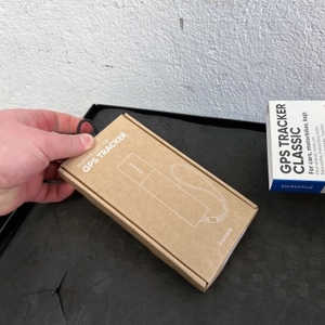 Der Invoxia GPS Tracker wird aus seiner Kartonverpackung gezogen. Die weiße Produktverpackung ist sichtbar.