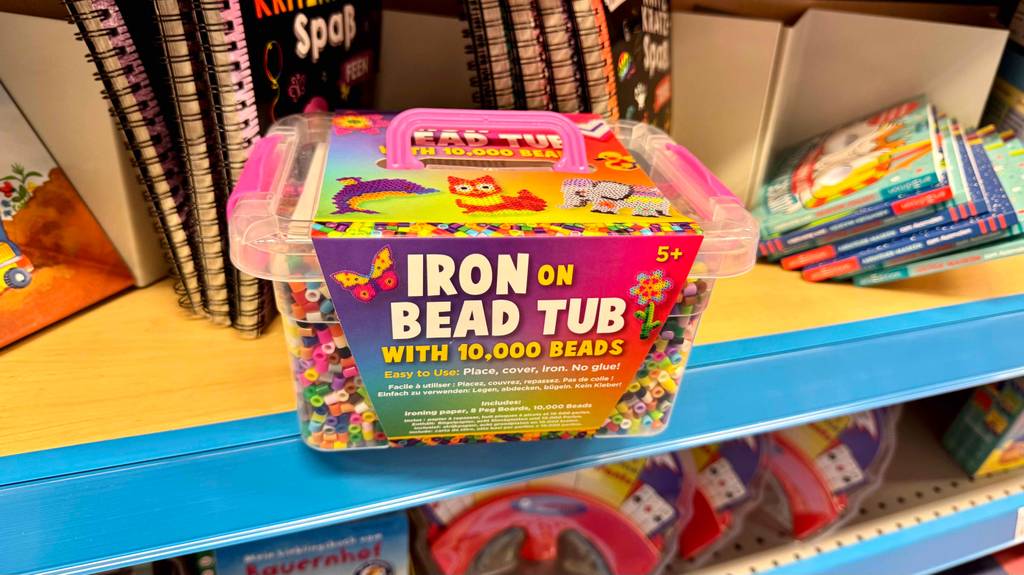 Die bunten, getesteten Bügelperlen in der transparenten 10 000‑Stück-Kunststoffbox befinden sich auf einem Verkaufsregal vor Kinderbüchern.