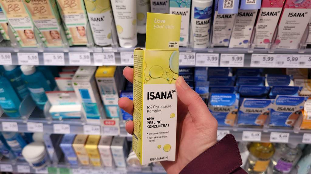 Hand hält eine weiß-gelbe Verpackung mit Isana Fruchtsäurepeeling im Test vor einem Verkaufsregal mit Hautpflegeprodukten.