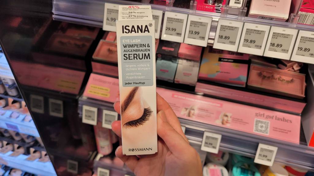 Eine Hand hält die Originalverpackung von getestetem Wimpernserum vor einem Kosmetikregal.