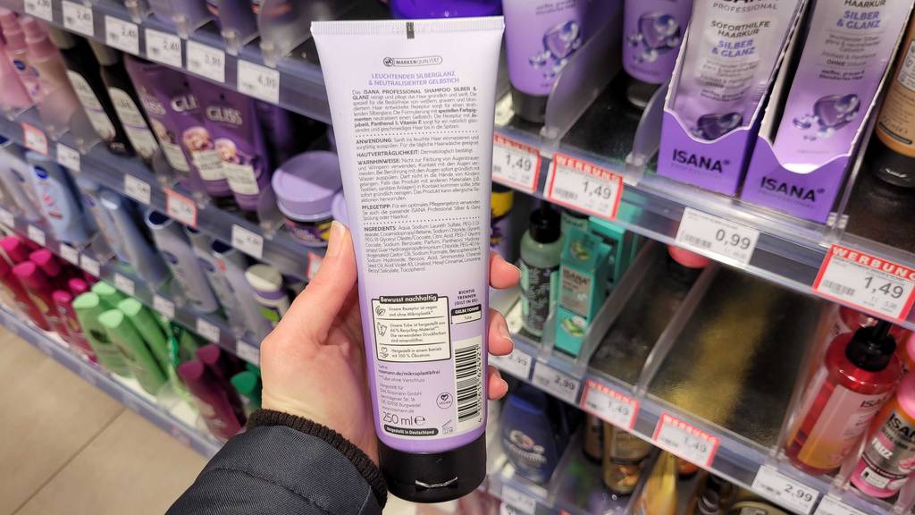 Hand hält eine Tube Isana Silbershampoo im Test, mit Fokus auf die rückwärtige Produktbeschreiubung, vor einem Ausstellungsregal mit Haarprodukten.