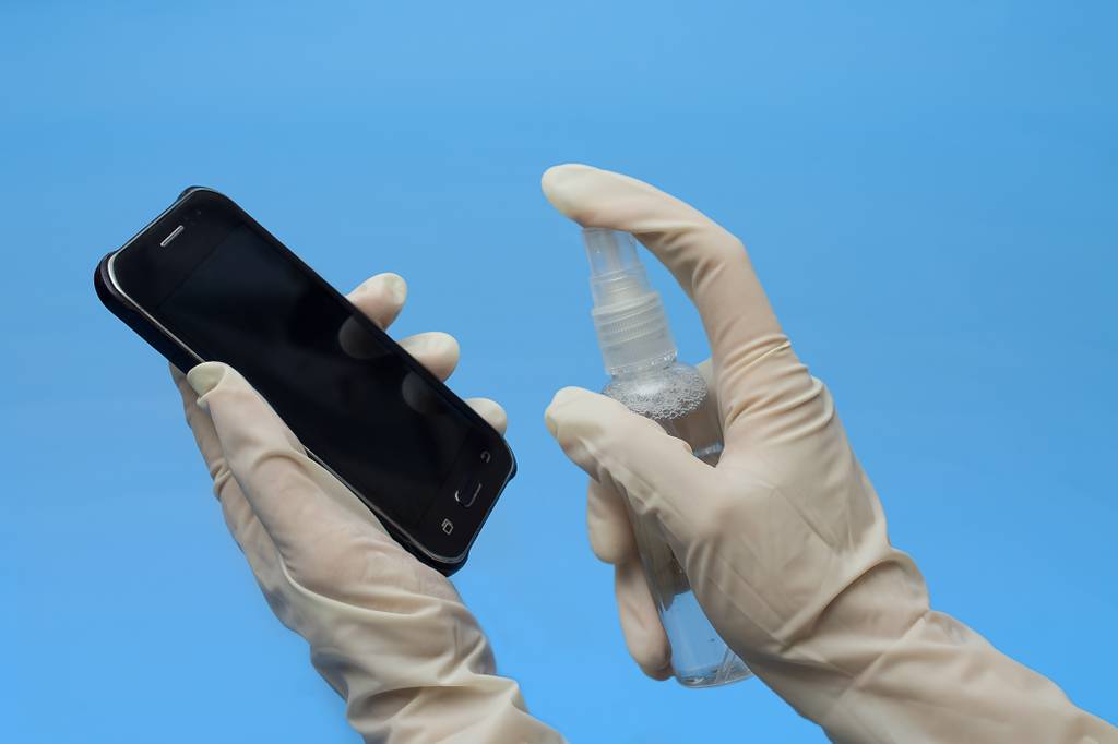 Eine Person trägt Schutzhandschuhe und reinigt ein Handy mit einer Isopropanol-Sprühflasche. 