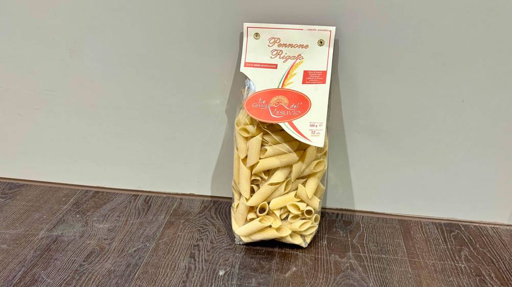 Vor einer grauen Wand steht die getestete italienische Pasta auf dem Holzboden.