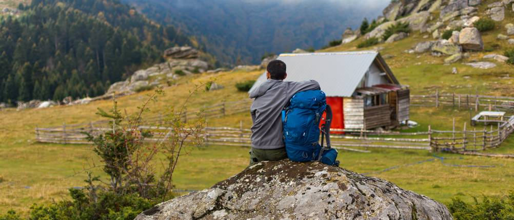 Jack-Wolfskin-Rucksack-Test: Mann, der mit seinem Rucksack neben sich auf einem großen Stein sitzt und auf ein Bergpanorama samt Hütte blickt